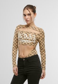 Bodysuit court à manches longues de couleur beige, décoré d'un motif logo noir et avec des côtés découpés. Fabriqué en tissu extensible et lisse. Pantalons noirs visibles.