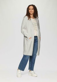 Jasnoszary długi kardigan z otwartym przodem, ribbowanym dołem i dwoma bocznymi kieszeniami, w połączeniu z kremowym T-shirtem i niebieskimi dżinsami.
