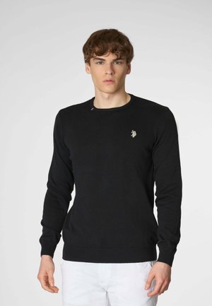 Un giovane uomo si trova in piedi contro uno sfondo semplice, indossando un maglione nero aderente con un logo discreto. Ha i capelli corti e ricci e un'espressione neutra.