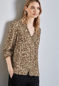 Blusa a fantasia marrone e beige con scollatura a V, maniche lunghe e chiusura con bottoni, abbinata a pantaloni neri. Tessuto morbido con una textura fluida.