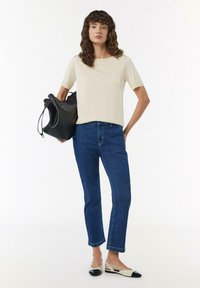 Beżowa bluzka z krótkim rękawem, niebieskie dopasowane jeansy i czarno-białe buty na odkrytej pięcie. Modelka trzyma czarną skórzaną torebkę. Prosty, casualowy strój.