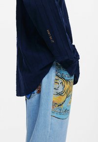 Marinebluse med tekstur og lodrette striber, der har "DON'T DO IT" broderi på ærmet; parret med blå jeans, der viser et farverigt tigermotiv.