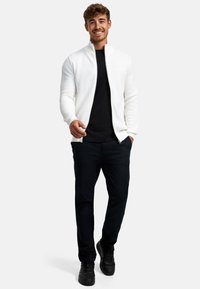 Chaqueta blanca texturizada con cremallera sobre una camiseta negra, combinada con pantalones negros y zapatos negros, en un modelo que está de pie contra un fondo liso.
