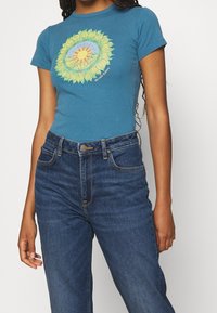 Teal cotton T-shirt featuring a colorful sunflower graphic in the center, paired with high-waisted blue denim jeans, showcasing classic styling.