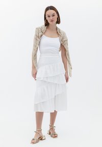 Witte gelaagde rok met ruches, gecombineerd met een witte tanktop en een beige gebreide sjaal. Sandalen met gouden accenten maken de outfit compleet.