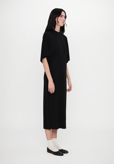Robe noire midi avec des manches courtes évasées, un col rond et une texture lisse et douce. Portée avec des chaussettes blanches et des ballerines noires.