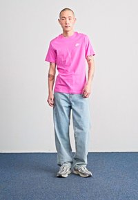 Rožinė trumpomis rankovėmis marškinėliai su baltu logotipu, derinami su šviesiai mėlynomis laisvais džinsais ir pilkais sportbačiais, stovint ant mėlynos kilimo fono.