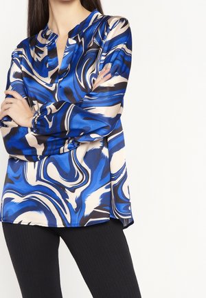 Žena v modré, černé a bílé abstraktně vzorované blůze s dlouhými rukávy, doplněné černými texturovanými kalhotami, s rukama zkříženýma na prsou.