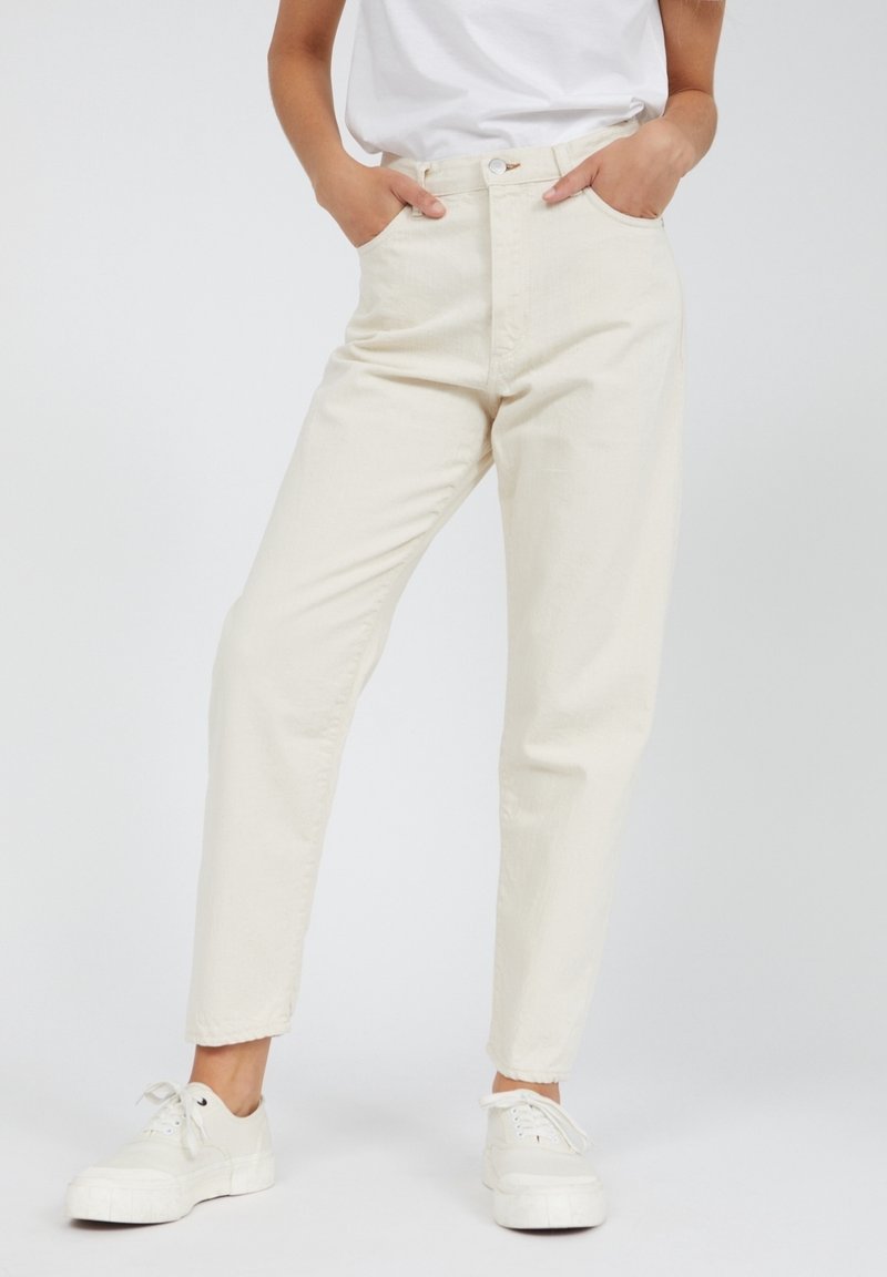 Personne portant un jean droit taille haute de couleur blanc cassé, les mains dans les poches, et des baskets blanches à lacets sur un fond uni.