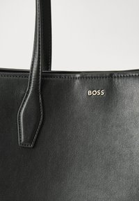 Crna kožna torba s glatkom teksturom, koja ima dva ručka i metalni logo "BOSS" sprijeda. Minimalistički dizajn.