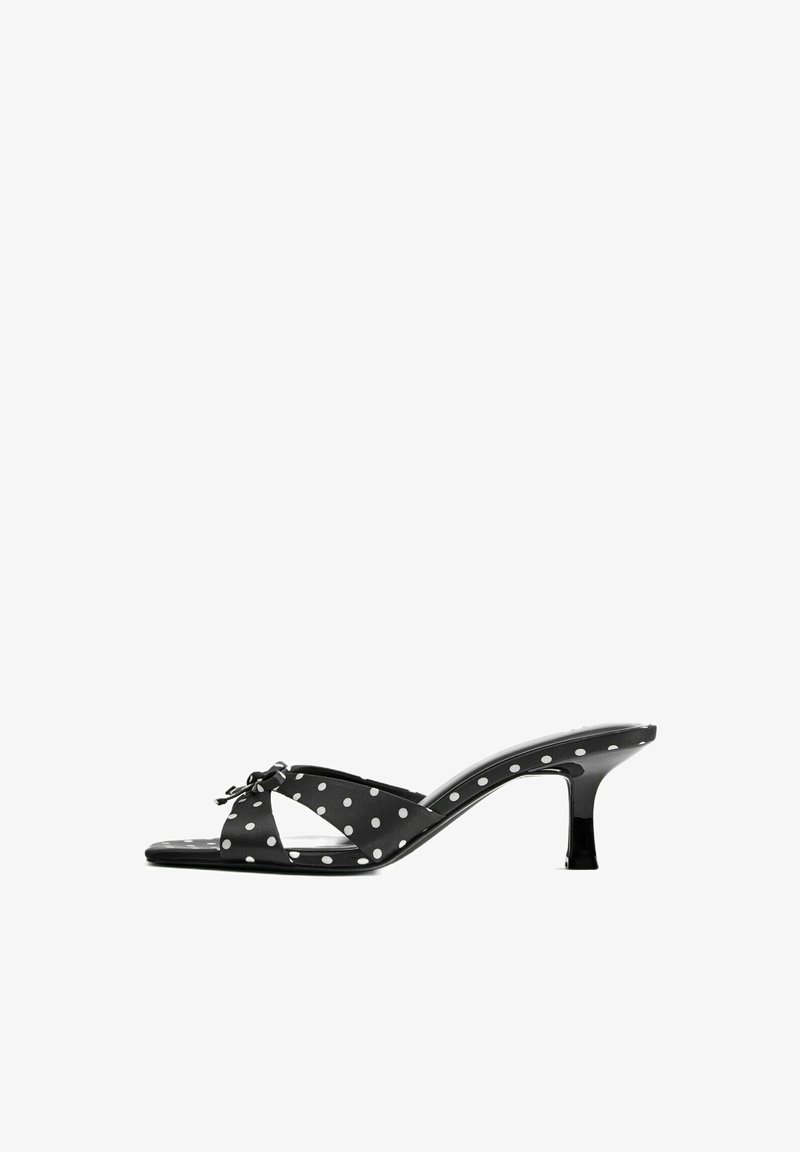 Sandale mule noire à bout ouvert avec des pois blancs, talon bas courbé et un petit nœud sur les brides croisées.