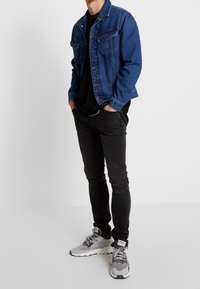 Colete de denim azul oscuro con bolsillos en el pecho, llevado sobre una camisa negra, combinado con jeans negros ajustados y zapatillas grises.