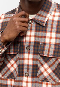 Košile s kostkovaným vzorem z texturované látky; má hnědé, modré a oranžové kostky, dvě přední kapsy a zapínání na knoflíky.
