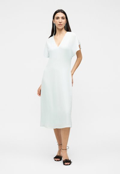 Γυναίκα με μακριά σκούρα μαλλιά που φοράει ένα λευκό midi φόρεμα με V-λαιμόκοψη και κοντά μανίκια και μαύρα ψηλοτάκουνα σανδάλια με φτερά, που στέκεται.