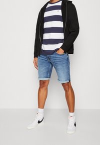 Sudadera negra con cremallera, camiseta a rayas en azul marino y blanco, pantalones cortos de mezclilla azul claro y zapatillas altas blancas de Nike con detalles en negro.