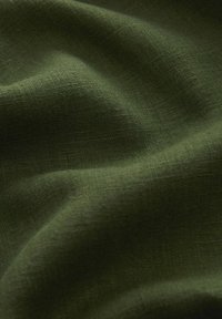 Zbliżenie na pomarszczony oliwkowy materiał z widoczną teksturą tkaniny oraz miękkimi fałdami tworzącymi wzory światła i cienia.
