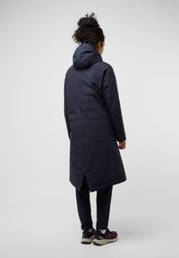 Dolg navy puhov coat s kapuco, ki ima razcepljen zadnji rob za enostavno gibanje in gladko, teksturirano blago.