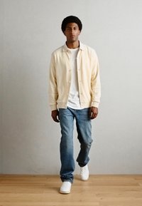 Cardigan giallo chiaro con bottoni, t-shirt bianca, jeans blu e sneakers bianche. Il pavimento è in legno, con uno sfondo grigio chiaro.