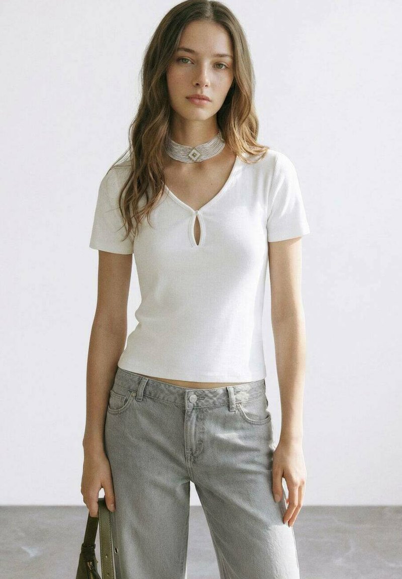 Jeune femme aux longs cheveux bruns portant un haut blanc à manches courtes, un jean gris et un collier ras du cou argenté, tenant un sac à main vert.