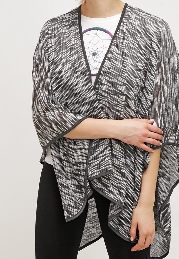 Personne portant un châle à motifs noir et blanc sur un t-shirt blanc imprimé d'un attrape-rêves et un pantalon noir, bras croisés à la taille.