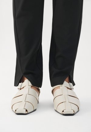 Personne portant un pantalon noir avec des ourlets fendus et des sandales fermées couleur crème avec des brides à boucle, debout sur une surface blanche.