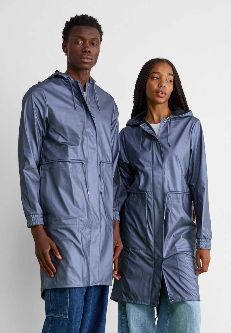 Vestes légères, imperméables, de couleur grise avec capuches. Poignets élastiques, poches frontales et coupe droite. Portées par deux modèles sur un fond neutre.