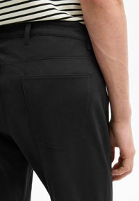 Zwarte katoenen broek met een achterzak, met een strakke uitstraling en subtiele textuur. Zichtbare stiksels bij de tailleband en de rand van de zak.
