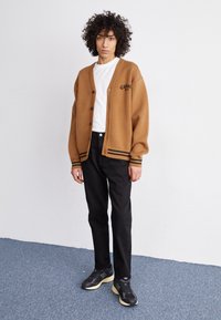 Brązowy, oversize'owy kardigan z czarnymi paskami na rękawach, czarnymi guzikami i logo. Noszony na białej koszulce, w zestawie z czarnymi spodniami i sneakersami.