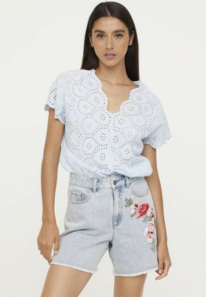 Blouse bleu clair en dentelle avec un col festonné, manches courtes, associée à un short en denim bleu clair avec des broderies florales sur la jambe gauche.