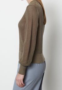 Dzianinowy sweter w oliwkowym zielonym kolorze, z teksturowanym otwartym splotem, długimi bufiaste rękawami i dopasowaną talią. Noszony z jasnoniebieskimi spodniami.