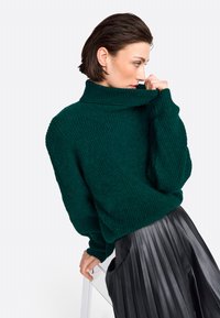 Grüner Strickpullover mit Rollkragen und strukturiertem Finish, kombiniert mit einem plissierten schwarzen Lederrock. Der Schnitt ist locker mit gesenkten Schultern.