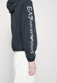 Svart nylonjacka med huva, featuring vit "EA7 EMPORIO ARMANI" text längs vänster ärm. Elastiska muddar och midja.