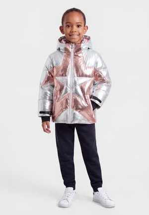 Enfant souriant portant une doudoune argentée et rose doré à motif d’étoiles, un pantalon noir et des baskets blanches, debout devant un fond blanc.
