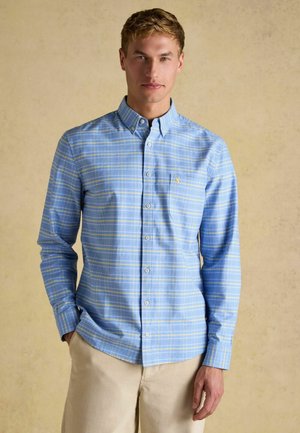 Mann trägt hellblau kariertes Hemd mit Knöpfen und beige Hose, steht vor schlichtem beigem Hintergrund mit einer Hand in der Tasche.