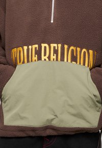 Polaire marron avec col zippé, dotée d'une poche avant vert olive et de l'inscription "TRUE RELIGION" brodée en doré au-dessus de la poche.