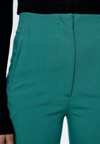 Teal hoge taillebroeken met een gladde textuur, voorzien van voorplooien en zijzakken, gecombineerd met een zwarte geribbelde blouse met lange mouwen.