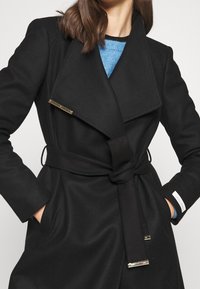 Zwarte wollen jas met een brede kraag en tailleband. Heeft gouden accenten en twee zijzakken. Soepele textuur en getailleerde pasvorm.