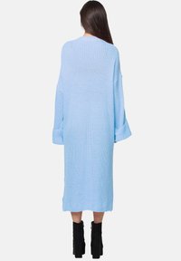 Abito lavorato a maglia di colore azzurro chiaro, dalla vestibilità ampia, con maniche lunghe e polsini arrotolati, che arriva a metà polpaccio. Presenta un motivo testurizzato. Sotto, stivali neri.