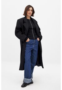 Svart överdimensionerad kappa lagd över en ribbad svart topp med knappar, i kombination med vida blå jeans och svarta skor. En enkel bakgrund.