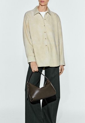 Beige suède overhemdjack met knopen, gecombineerd met donkergroene broek; ze houdt een donkerbruine leren handtas met een gebogen ontwerp vast.