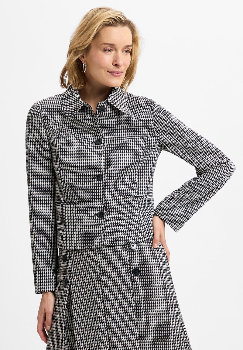 Houndstooth-patterned jacket featuring a collar, black buttons, and two front pockets. Textured fabric with a fitted silhouette.