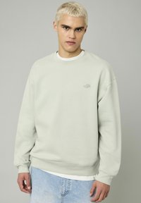 Sweatshirt vert menthe avec un col rond, des épaules tombantes et des poignets côtelés. Il présente un petit logo sur la poitrine et est associé à un jean en denim clair.