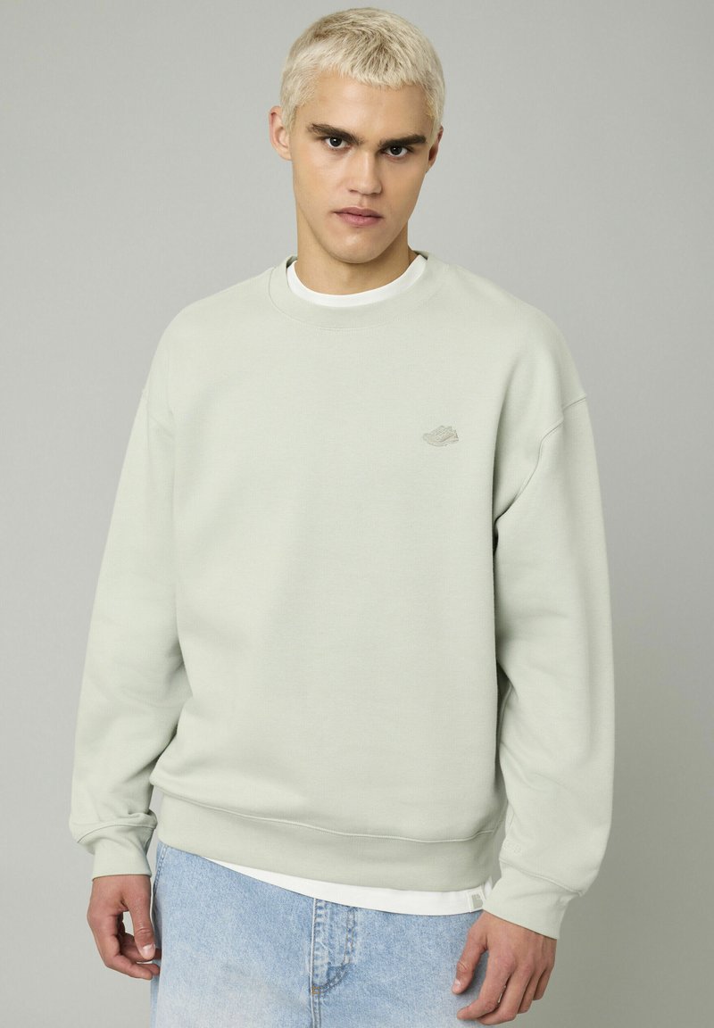 Sweatshirt vert menthe avec un col rond, des épaules tombantes et des poignets côtelés. Il présente un petit logo sur la poitrine et est associé à un jean en denim clair.