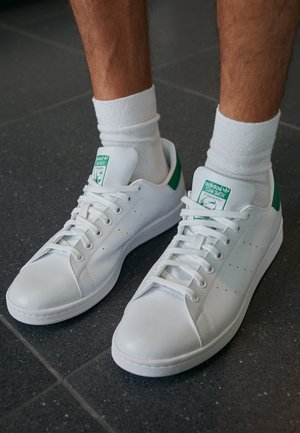 Personne portant des baskets blanches Adidas Stan Smith avec des languettes vertes au talon et des chaussettes blanches, debout sur un sol carrelé foncé.
