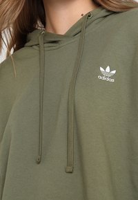 Olive green hooded sweatshirt with drawstrings. Features a white Adidas logo on the chest. Material appears soft with a smooth texture.