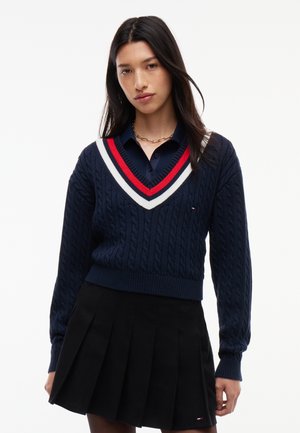 Kvinde med langt mørkt hår iført navy kabelstrikket sweater med røde og hvide striber ved halsudskæringen over sort plisseret nederdel mod hvid baggrund.
