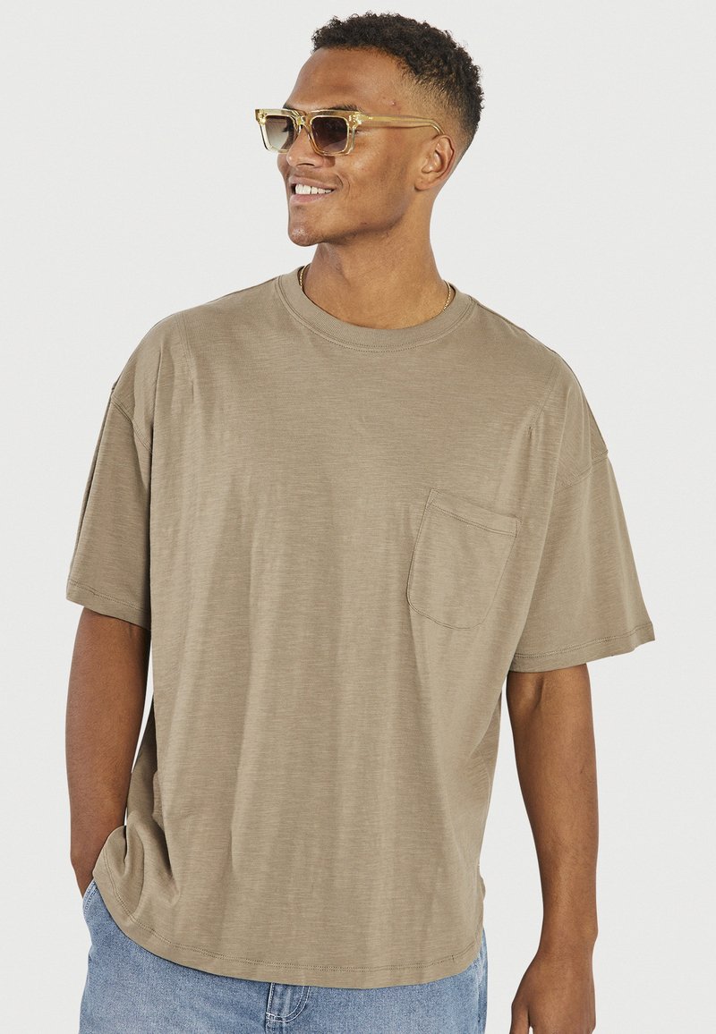Beige short-sleeve t-shirt made of soft fabric, featuring a chest pocket and a crew neckline. Paired with light blue denim shorts.