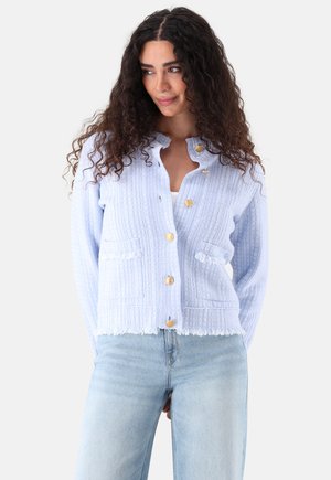 Donna che indossa un cardigan azzurro chiaro con bottoni dorati e dettagli di frange, abbinato a jeans azzurri, che guarda di lato.