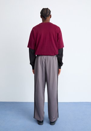 Homme aux cheveux tressés, debout face à un mur blanc, portant une chemise bordeaux à manches courtes sur un haut noir à manches longues et un pantalon gris à jambes larges.