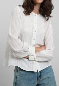Blusa bianca a maniche lunghe con chiusura a bottoni, caratterizzata da una texture in pizzo sul davanti e tessuto a righe sulle maniche. Abbinata a jeans blu.